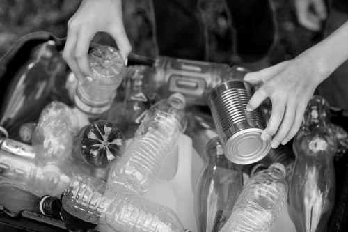 Close-up of a person's hands sorting and collecting various discarded plastic bottles and cans, including clear and dark bottles, into a container or collection box. The collection showcases different shapes and sizes of empty, used beverage containers made of plastic and metal, some with visible caps and labels. The background suggests an outdoor environment, possibly a waste collection or recycling activity, with the focus on the hands handling the waste materials. The scene emphasizes the act of waste sorting, relevant to rubbish removal services such as those provided by Commercial Waste Removal Honor Oak, operating in the local area around Honor Oak and the postcode region. The lighting appears natural, highlighting the textures of the plastic bottles and metallic cans, with the overall tone neutral and professional, suitable for illustrating waste management activities.
