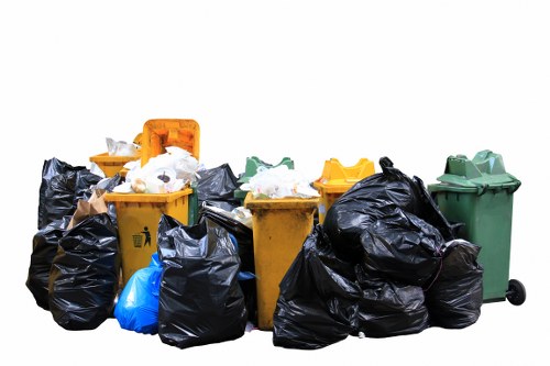 A collection of waste containers and trash bags placed outdoors on a paved or concrete surface, including several large black plastic rubbish bags filled with waste, some tied and others open, revealing mixed refuse inside. There are three wheelie bins in distinct colours: yellow, green, and another greyish-green, positioned behind and to the sides of the bags, with the green bin on the far right and the yellow bins closer to the center. The yellow bins are partially open, showing some discarded paper and plastic waste inside. The scene appears to be on a driveway or yard near a residential or commercial property in Honor Oak, with a plain white background emphasizing the rubbish and containers. The arrangement suggests an ongoing rubbish removal process, consistent with services offered by Commercial Waste Removal Honor Oak, typical of waste collection for local businesses or households in the area, possibly within the postal code region served by the company in London.