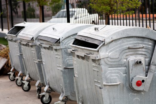 A row of four large, silver metal wheeled dumpsters lined up on a paved outdoor surface, likely a driveway or loading area in Honor Oak. The dumpsters are rectangular with rounded tops and are equipped with black lids, some of which are slightly open or tilted. Each container has visible handles and hinges along the sides, with one featuring a small red circular lock or latch on its front panel. The background shows a black iron fence, with trees and part of a street in the distance, indicating a residential or commercial environment in Southeast London. The overall scene emphasizes waste management infrastructure typical of rubbish removal services, with a focus on durable, weather-resistant metal containers used for collecting and transporting waste.